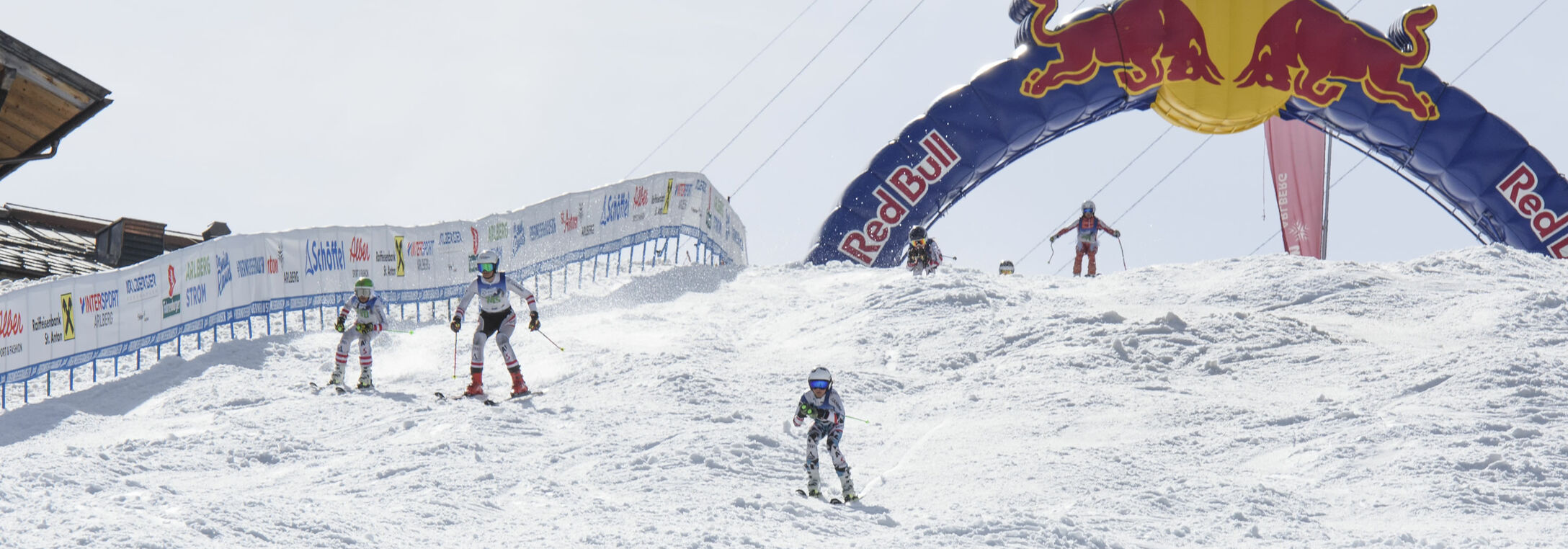 Der Weisse Rausch Mini Vier Kinder rasen auf Skiern dem Ziel in St. Anton am Arlberg entgegen. Sie durchfahren einen RedBull Bogen und bewältigen die buckelige Piste.