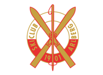 Logo des Skiclub Arlberg mit zwei gekreuzten roten Skiern und dem Schriftzug „Ski Club Arlberg 1901“. Es symbolisiert Tradition im alpinen Skisport.