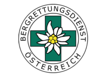 Logo der Bergrettung Österreich, weißes rundes Emblem mit doppeltem Rand, in der Mitte ein grünes Pluszeichen, verziert mit einer Edelweißblume.
